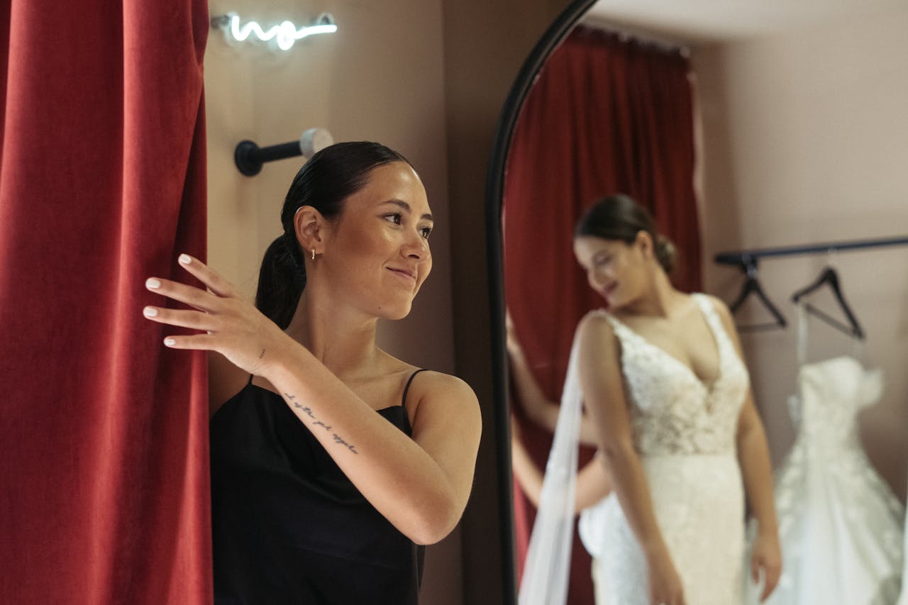Woman in fitting room with a wedding dress, smiling in front of a mirror with red curtain.